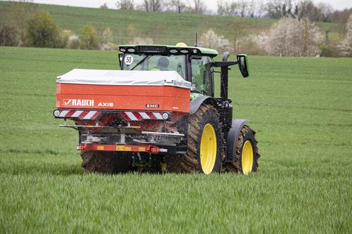 Ein landwirtschaftlicher Traktor fährt über ein grünes Feld und bringt mit einem Düngerstreuer Mineraldünger aus, der als kleine Partikel hinter dem Fahrzeug auf die Pflanzen verteilt wird. Im Hintergrund sind leicht hügelige Felder und blühende Bäume zu sehen.